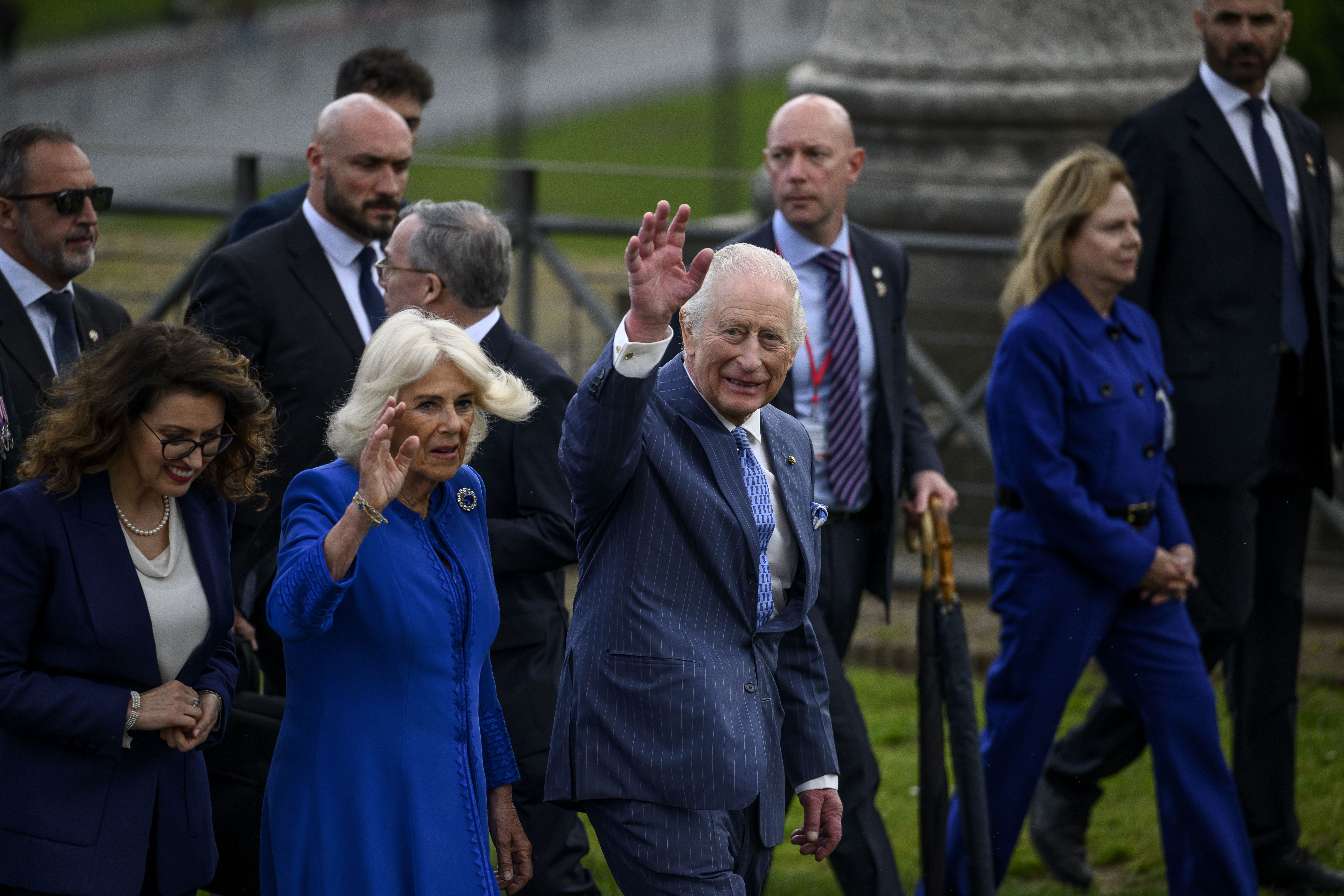 King Charles III and Queen Camilla share a wave with onlookers as they walk through a public space in Rome, flanked by a circle of suited security and aides. Charles, dressed sharply in a navy pinstripe suit, looks composed yet warm, while Camilla, radiant in cobalt blue, flashed a gentle smile.