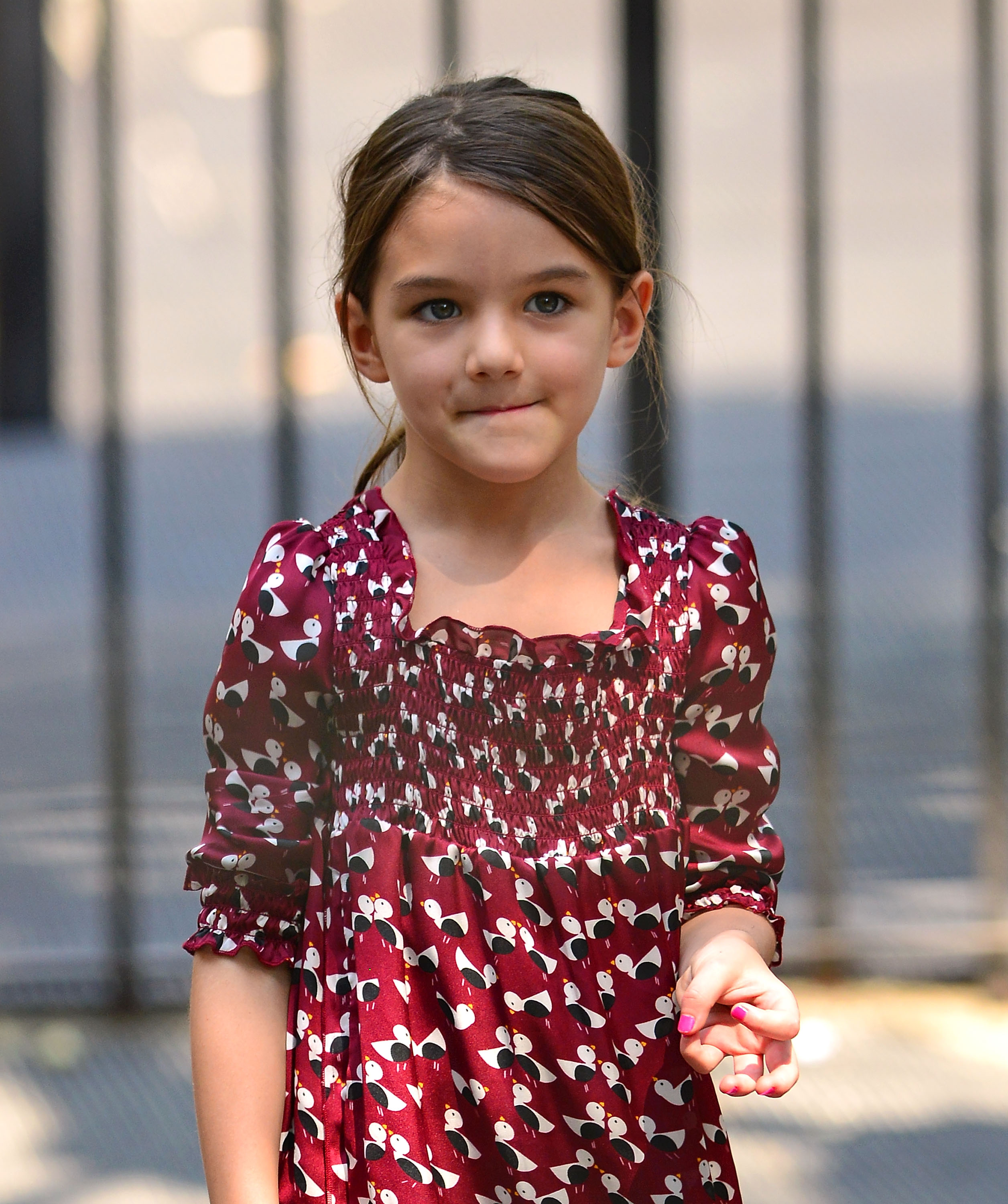 Bathed in soft daylight, young Suri gazes ahead with quiet confidence and a hint of curiosity. Her maroon dress, patterned with playful designs, adds a touch of charm to her poised demeanor. With her pink-painted nails and thoughtful expression, she embodies grace in simplicity.