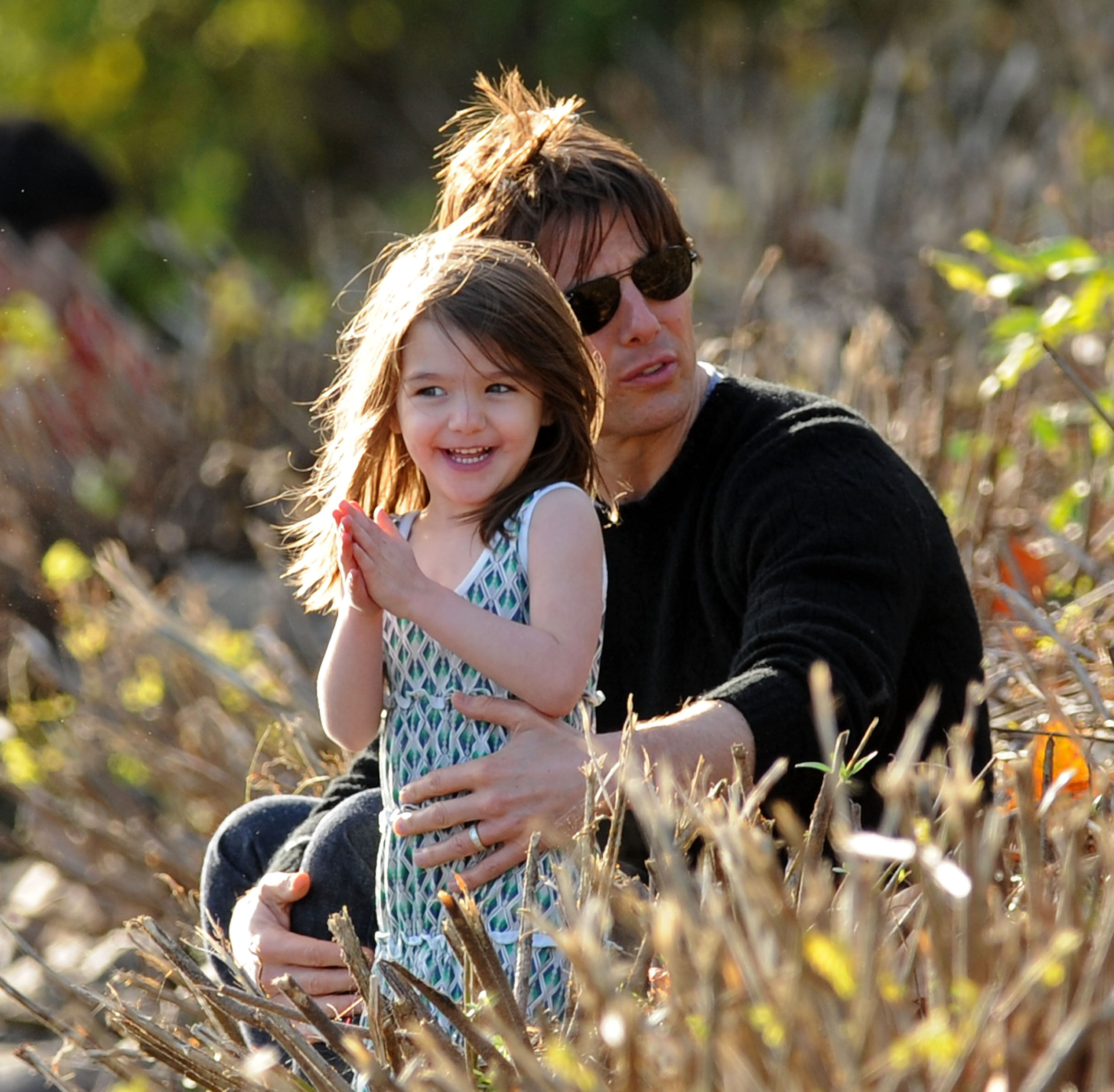Amid sunlit branches and golden hues, Tom Cruise sits protectively beside a gleeful Suri. His laughter lights up the moment, pure and unfiltered, as his steady presence anchors the joy. Together, they share a quiet, beautiful bond that outshines the world around them.