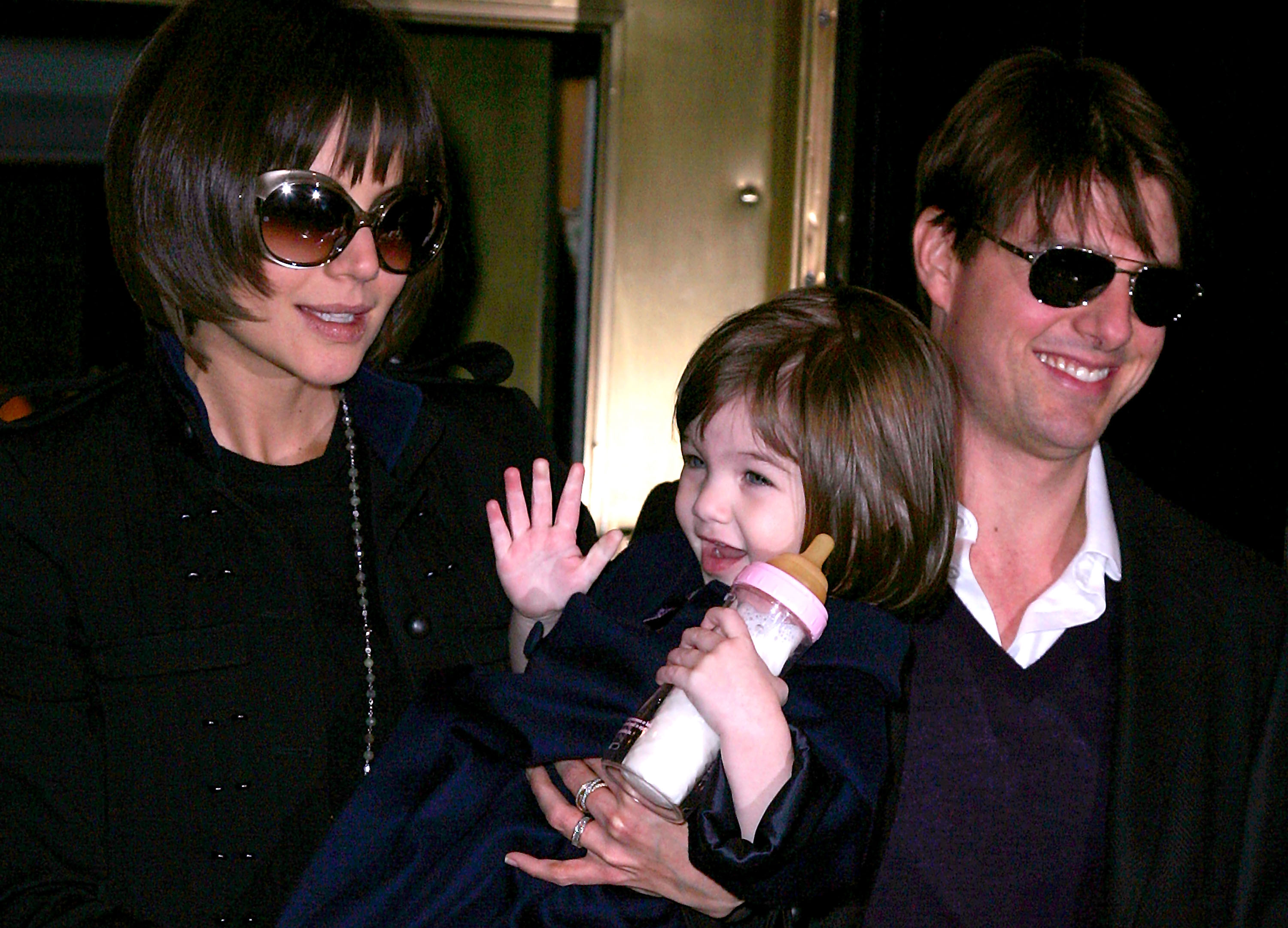 With matching shades and radiant smiles, Tom Cruise and Katie Holmes step out arm in arm, exuding effortless Hollywood cool. Little Suri, beaming with joy and clutching her bottle, waves sweetly at the crowd. The trio’s warmth and charm turn an ordinary outing into a picture-perfect family moment.