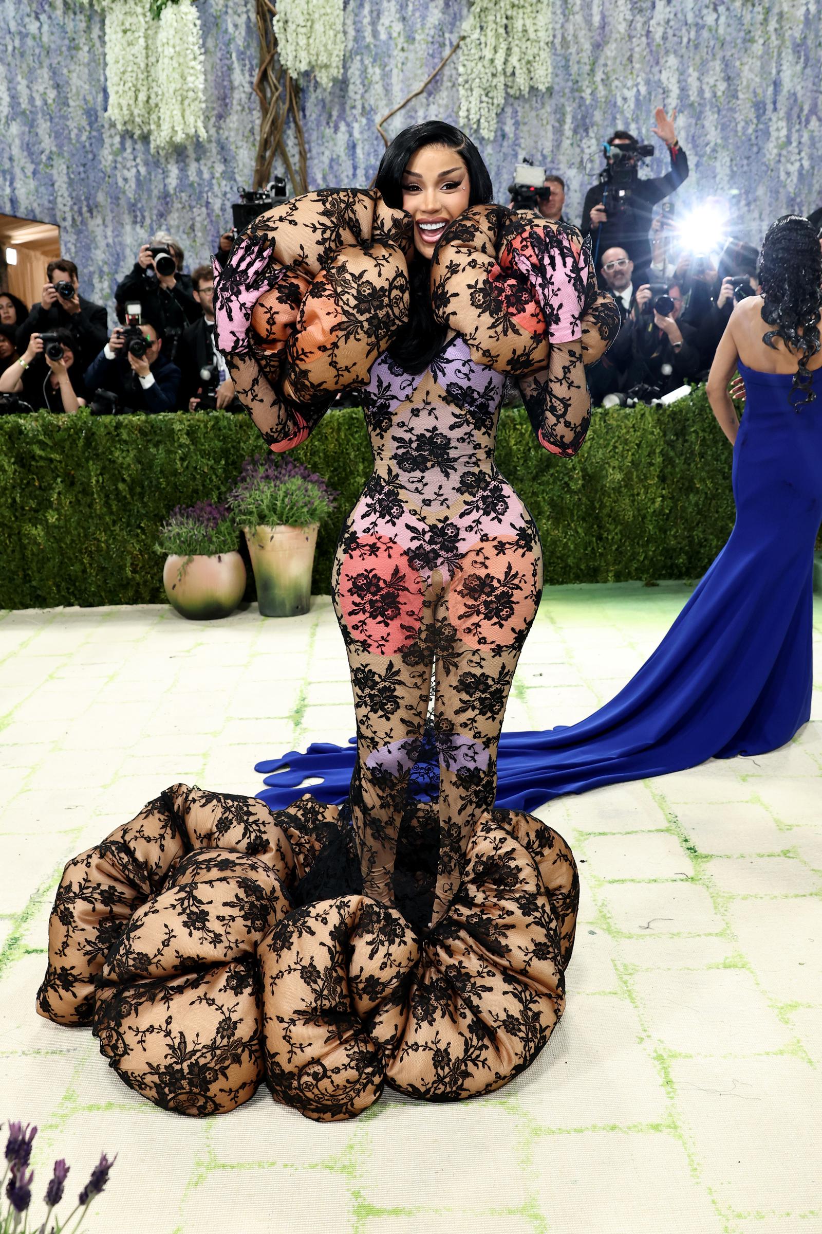 Cardi B beaming in a black lace floral catsuit over a multicoloured pink, orange and lavender base, with enormous inflated lace-covered balloon forms erupting from both shoulders and coiling into a massive sculptural heap at her feet.