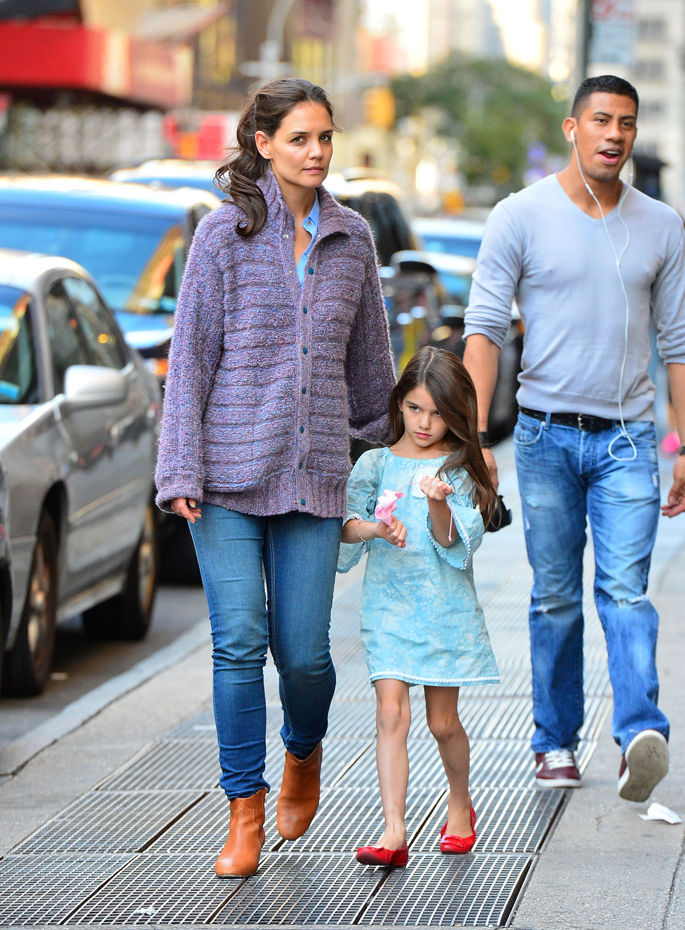 Katie Holmes strides through the city with calm confidence, her hand gently clasping young Suri’s. Dressed in a cozy sweater beside her daughter’s playful blue dress and red flats, the pair embody effortless style. Amid the city’s buzz, their quiet bond speaks louder than the noise around them.