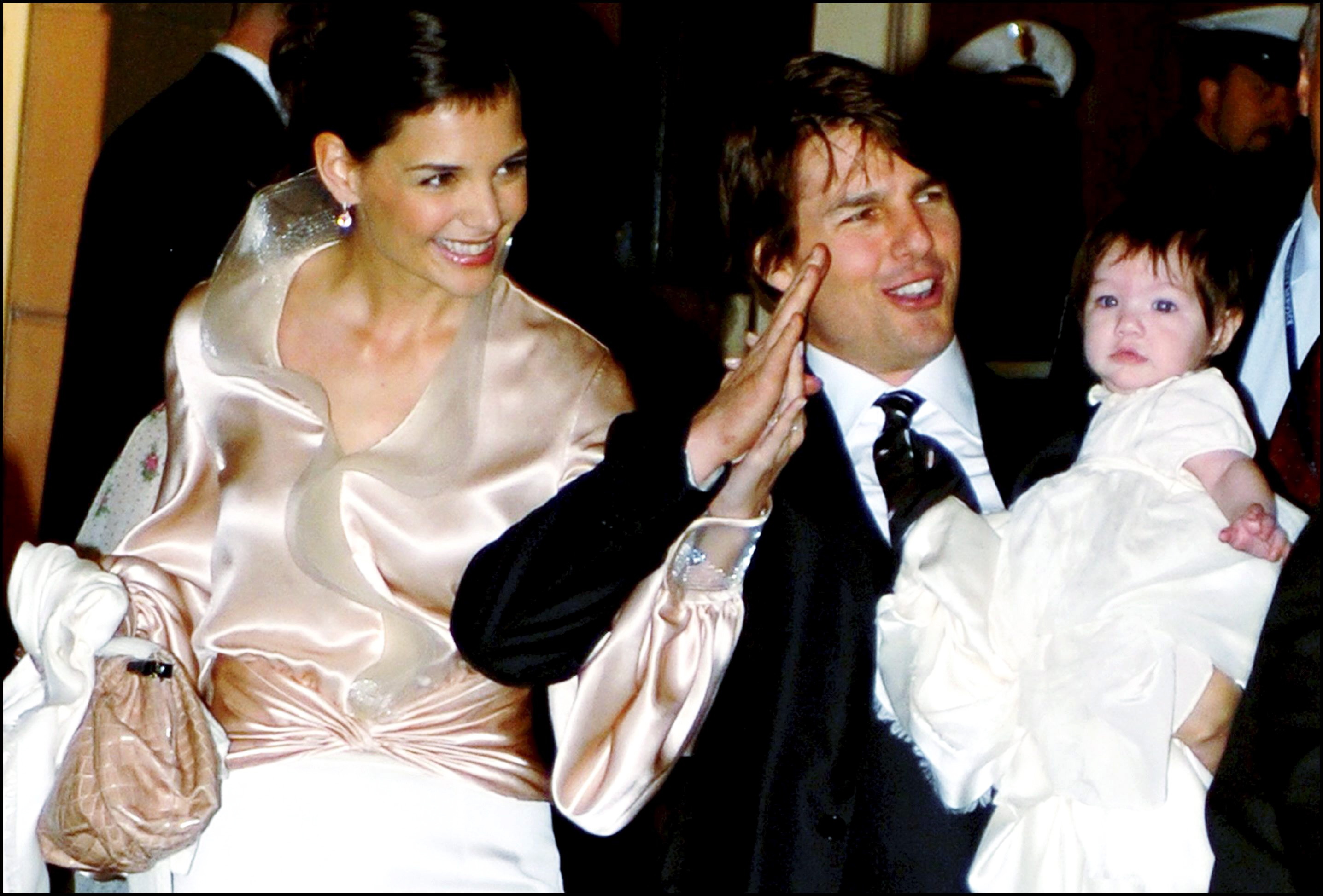 Dressed in elegance, Katie Holmes beams beside Tom Cruise, who proudly waves while cradling their baby daughter, Suri Cruise. The family radiates Hollywood glamor wrapped in warmth and affection. Under the flash of cameras, their smiles capture a rare, tender moment of love in the spotlight.