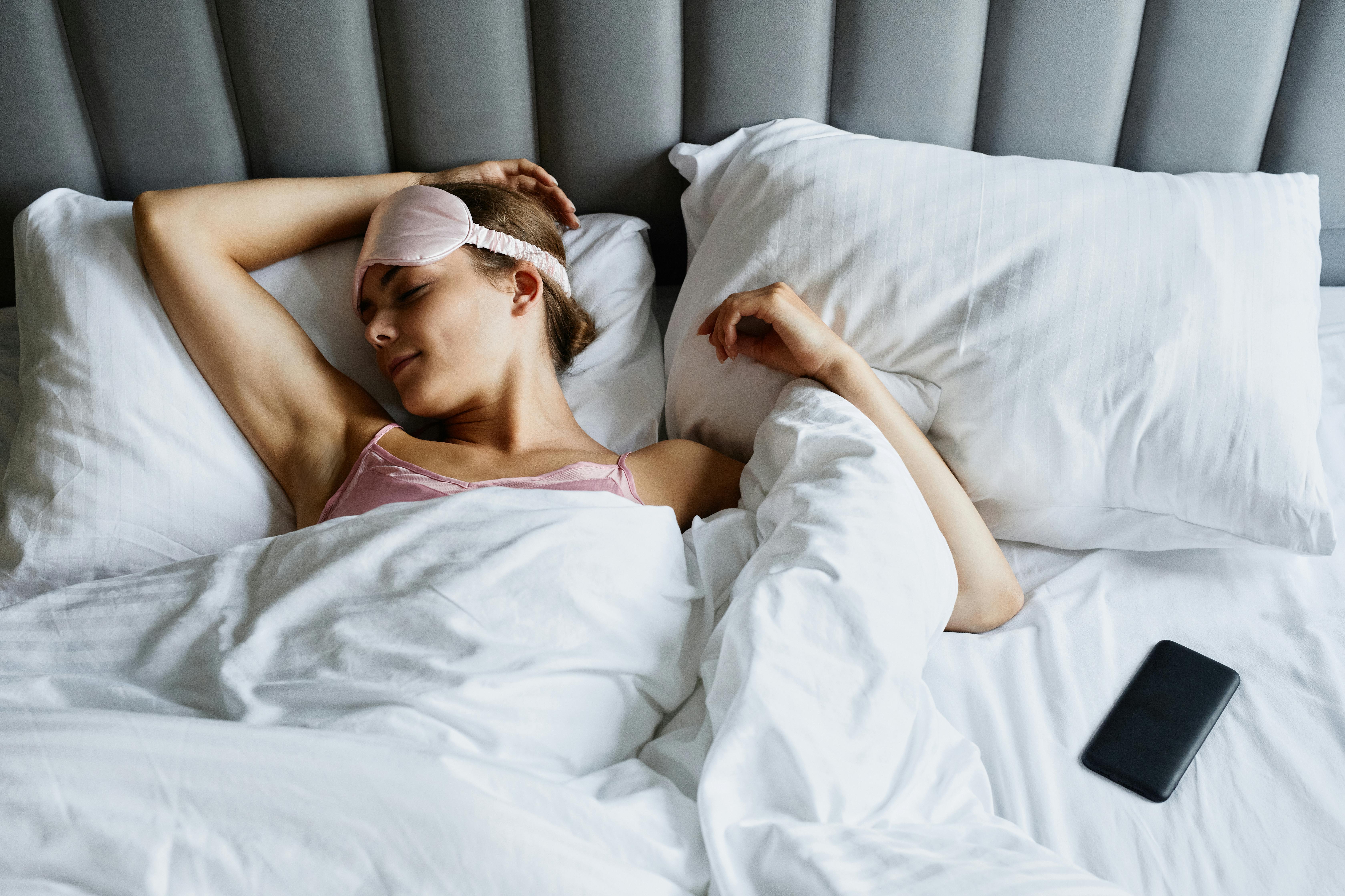 A woman sleeps peacefully in bed wearing a pink sleep mask, surrounded by white pillows and bedding. | Source: Pexels