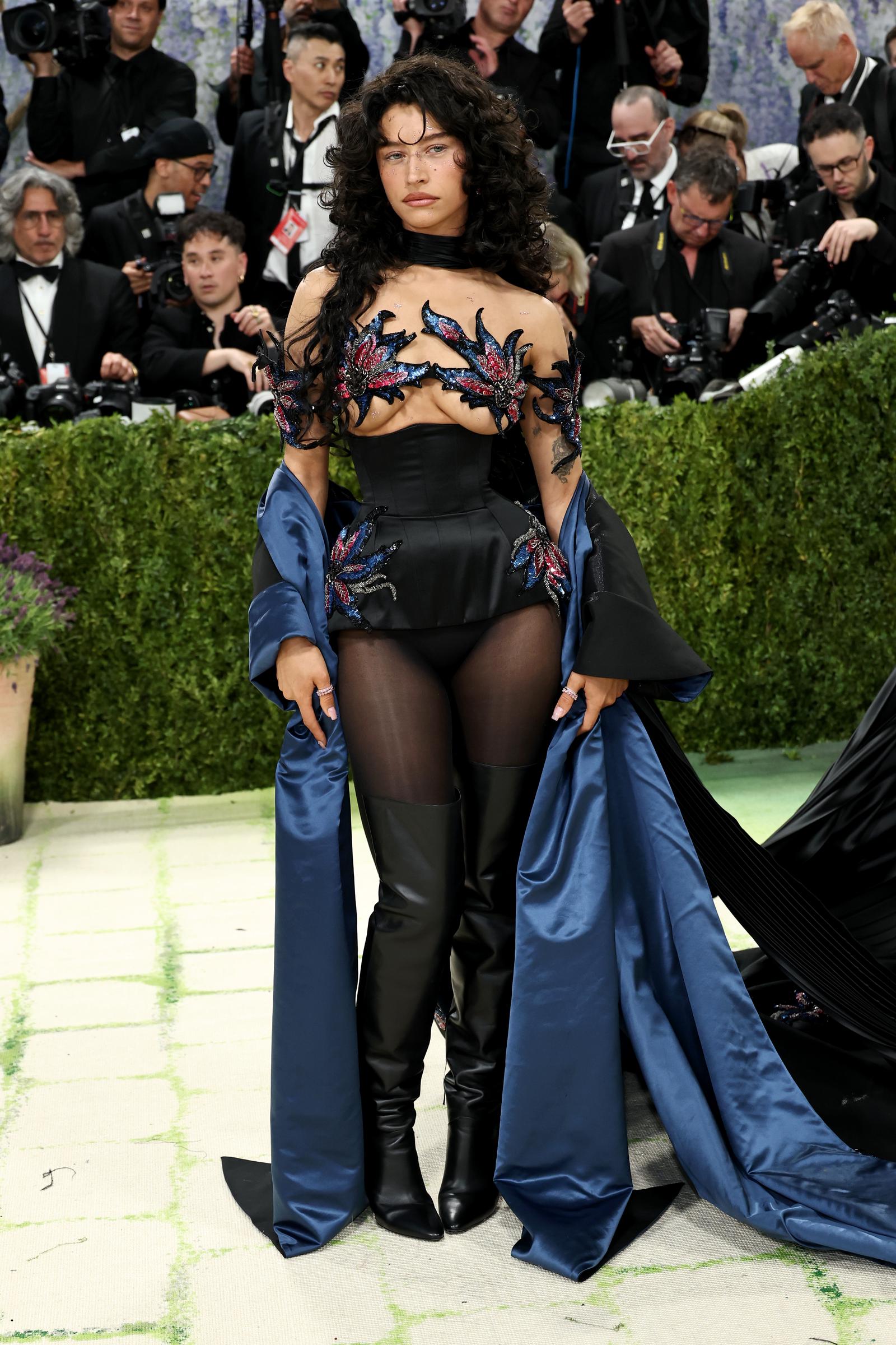 Odessa A'zion in a black corseted micro-skirt with sheer sleeves featuring red and blue sequinned floral appliqués, black thigh-high leather boots, sheer tights, and a dramatic royal blue and black satin train trailing behind her.