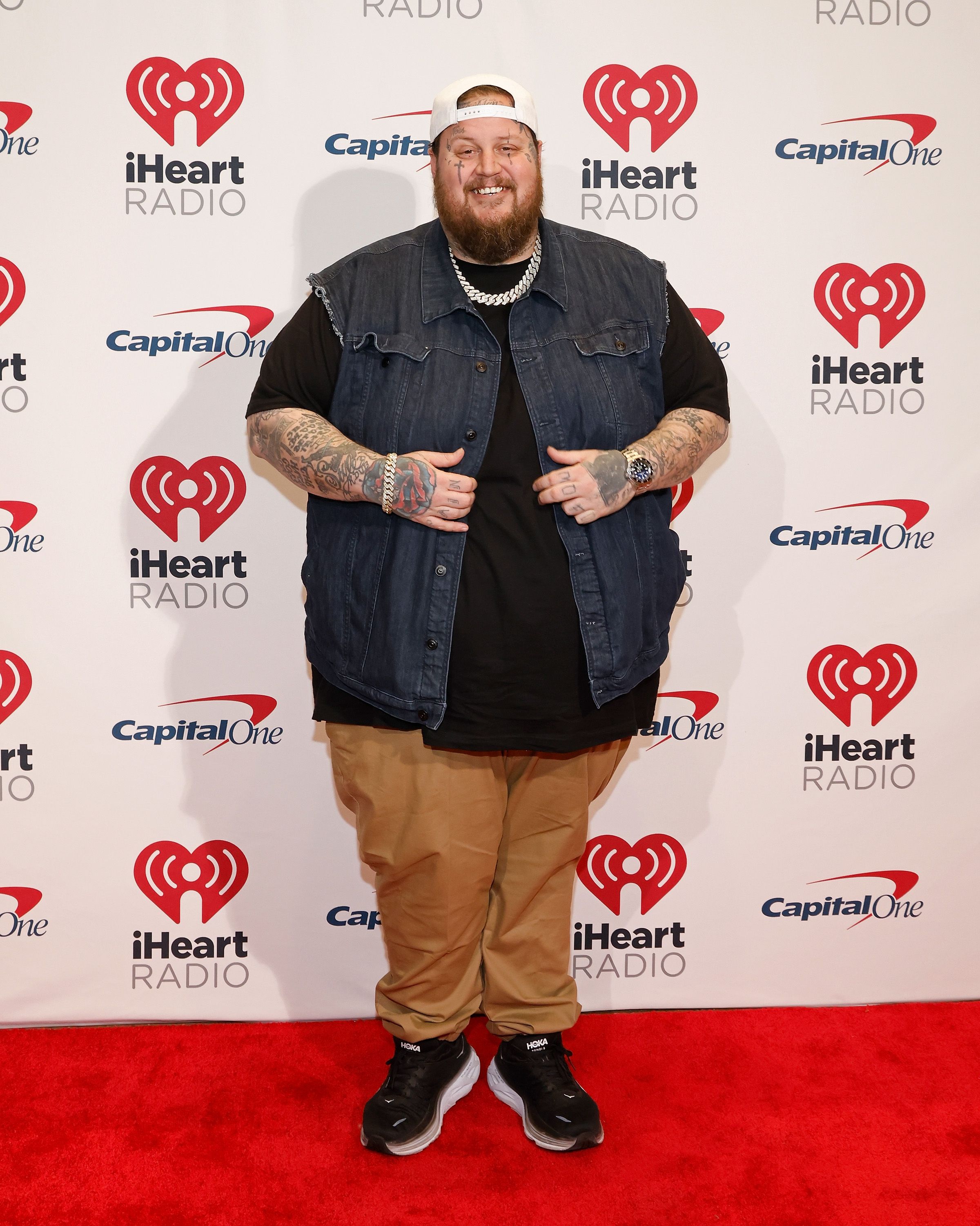 Jelly Roll poses on the red carpet at the 2023 iHeartRadio 106.1 KISS FM Jingle Ball at Dickies Arena on November 28, 2023, in Fort Worth, Texas, wearing a black shirt, brown pants, a denim vest, and a diamond chain necklace.