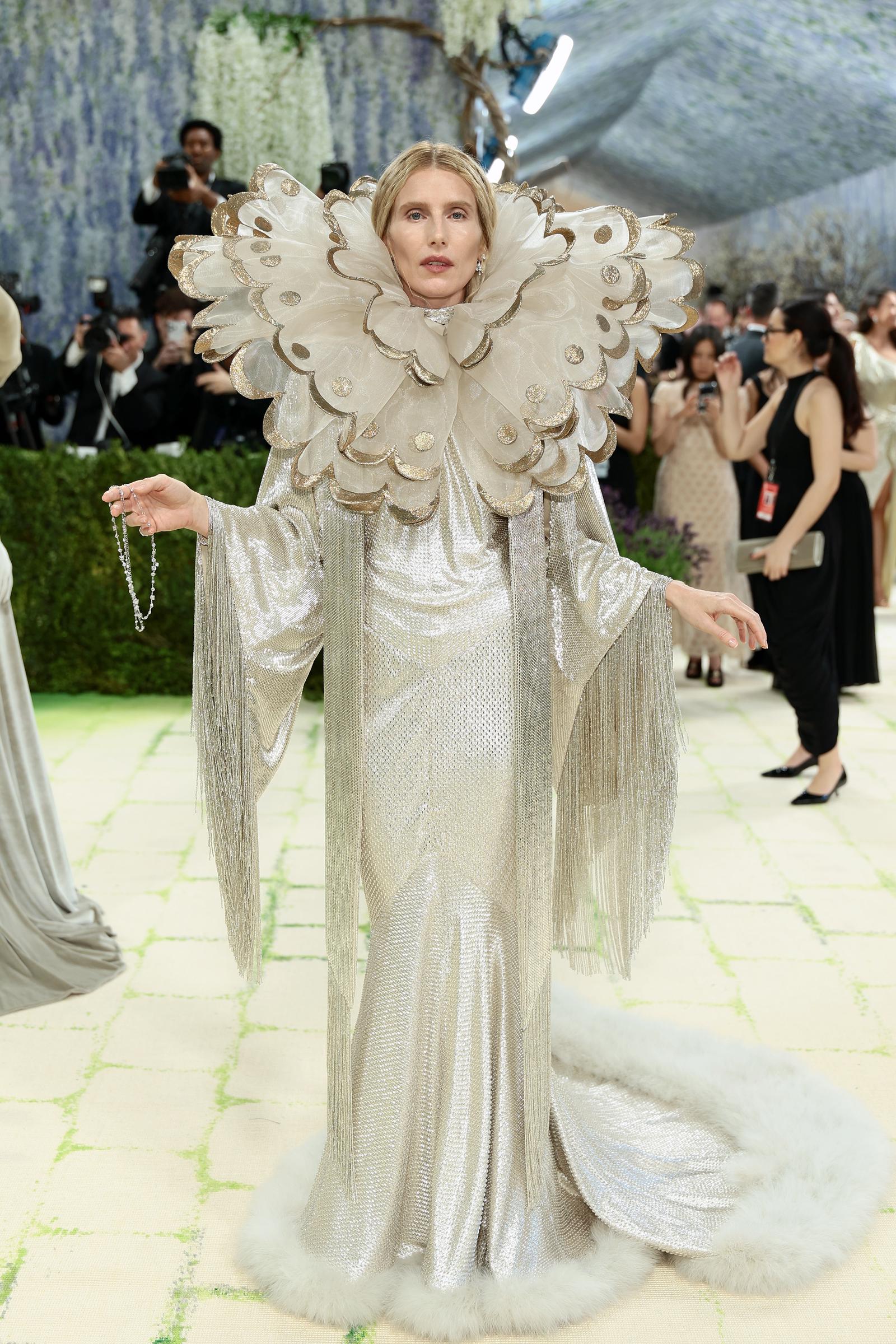 Dree Hemingway in a silver chainmail column gown with long fringe sleeves, a fluffy white feather hem, a dangling crystal bracelet, and an extraordinary oversized gold-tipped butterfly wing collar fanning out around her face like an Elizabethan ruff.