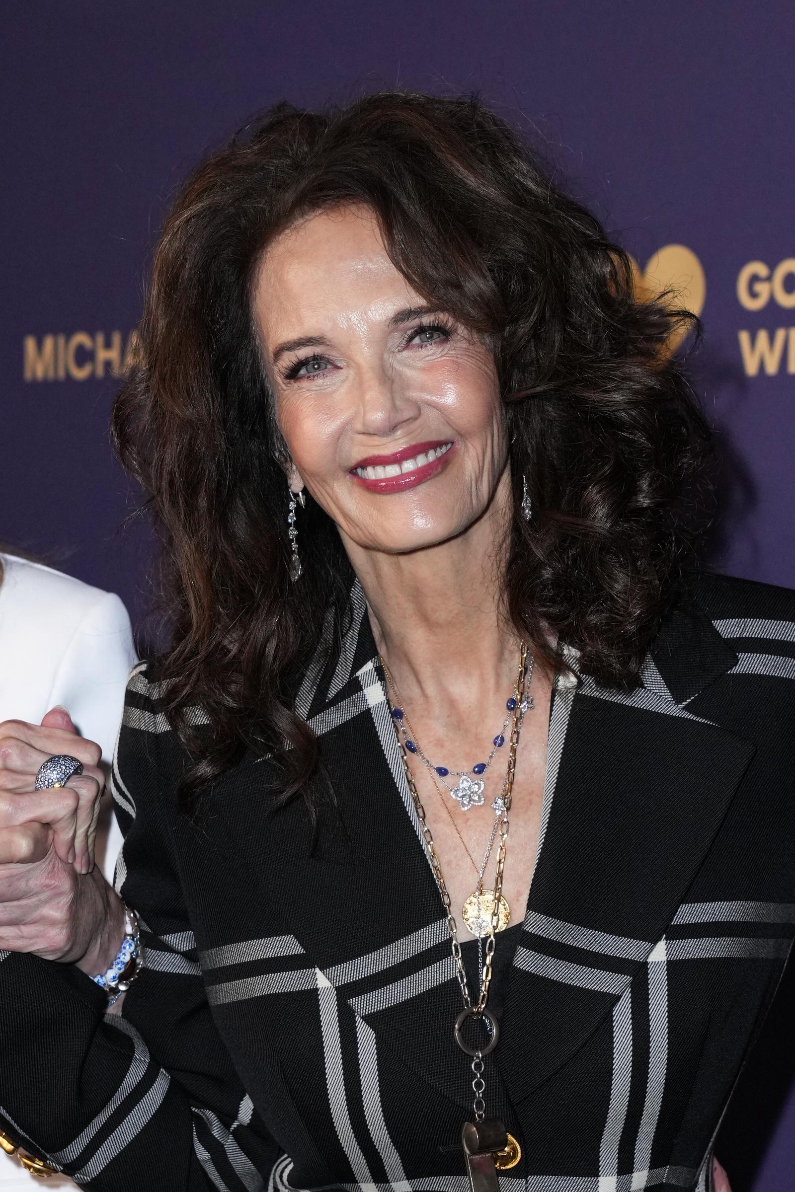 Lynda Carter flashes a radiant smile on the red carpet at the God’s Love We Deliver event, wearing a black plaid blazer with layered gold and gemstone necklaces. Her voluminous curls frame her face as she accessorizes with drop earrings and a bold pink lip, exuding warmth and star power.