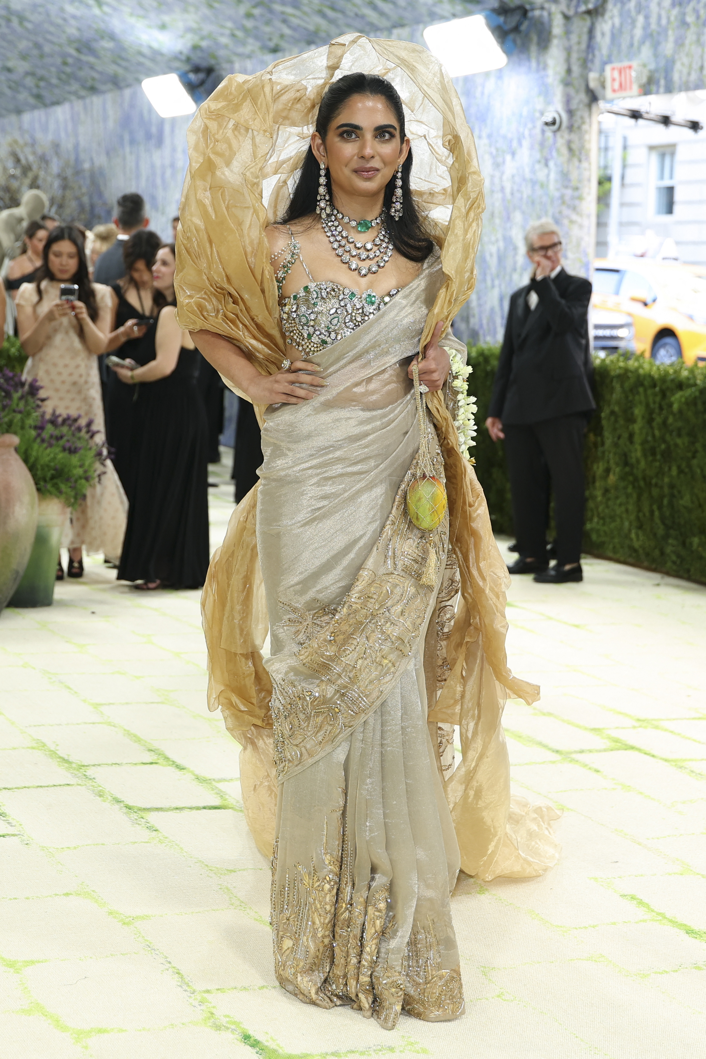 Isha Ambani in a silver and gold embroidered saree with intricate border detailing, a jewelled emerald and diamond bralet bodice, layered diamond and emerald necklaces, a voluminous crinkled gold organza dupatta forming a halo around her head, and a small mango held in a jasmine garland in her hand.