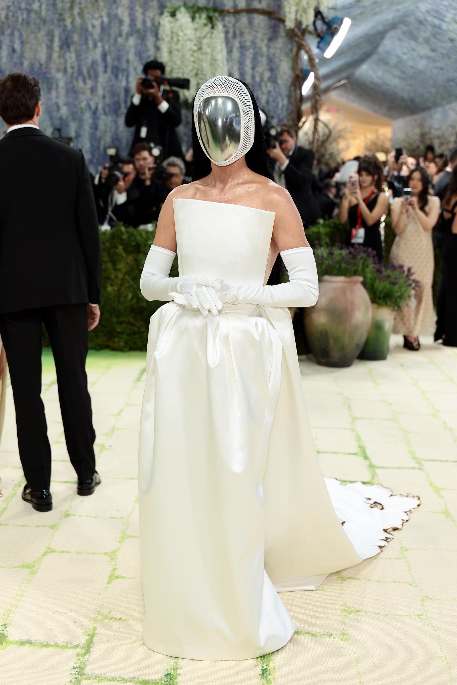 Katy Perry front-facing in a structured white strapless satin ballgown with a gathered fabric detail at the waist, white opera gloves, a scorched and frayed hem on the train, and a polished chrome dome visor encased in white mesh netting that fully obscures her face.