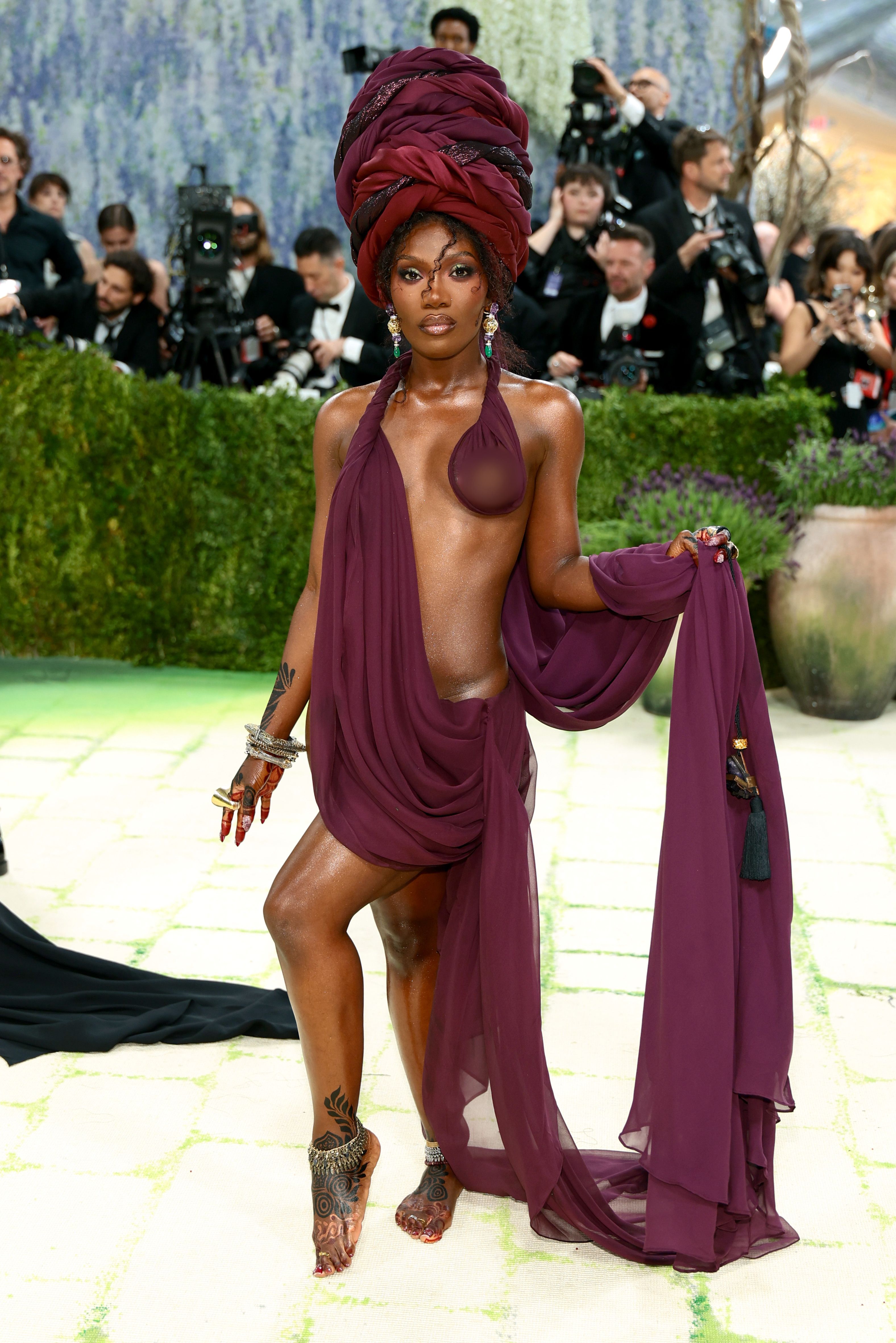 Doechii revealed on the Met carpet in a deep crimson draped halter with a towering sculptural headwrap, henna-tattooed bare feet adorned with silver anklets, stacked bracelets, red talon nails, and a tasselled fabric train held aloft in one hand.