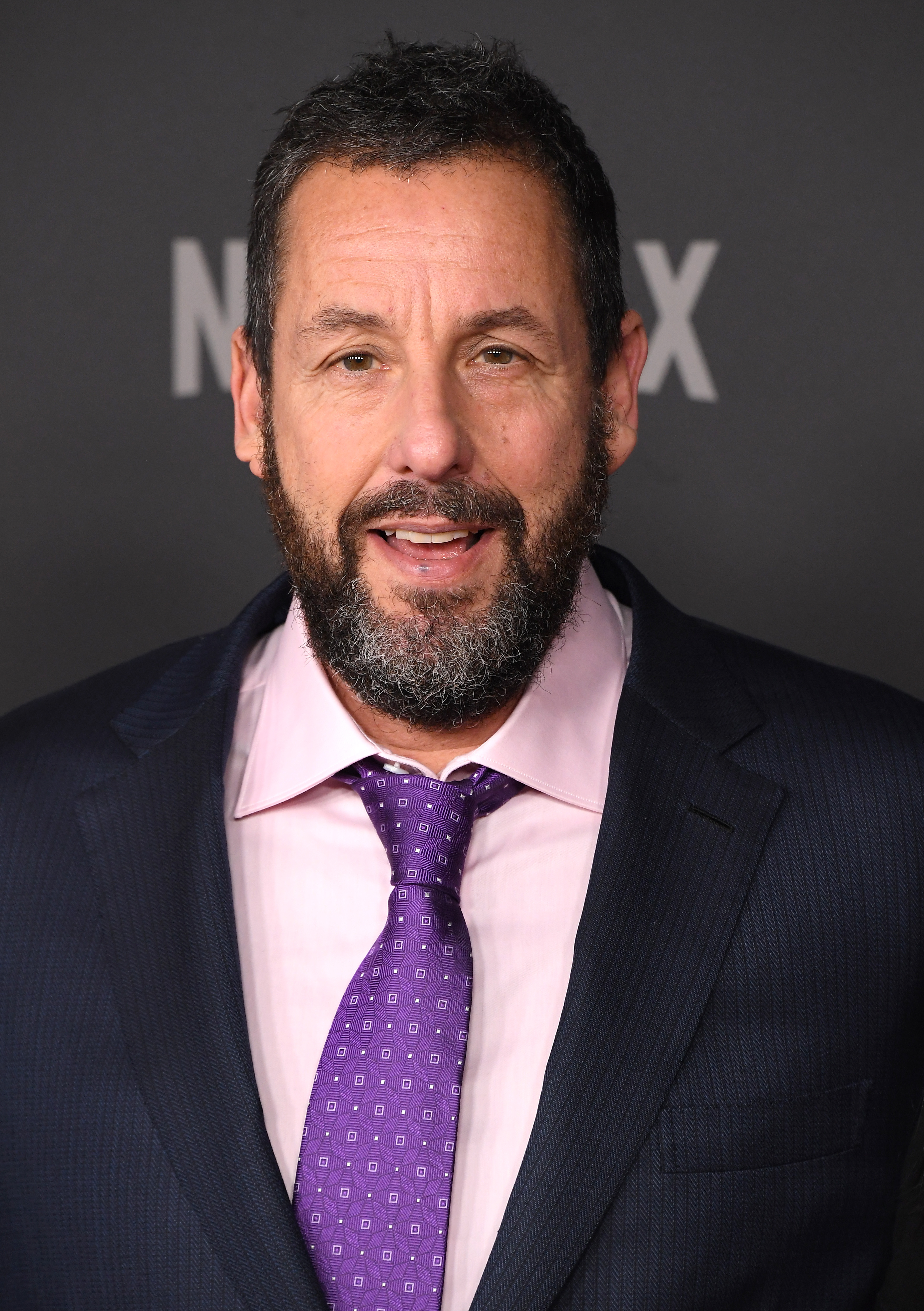 A close-up shot captures Adam Sandler at the premiere of Netflix’s "Jay Kelly." He wears a slight smile as the camera focuses on his graying beard and hair, highlighting his timeless charm.