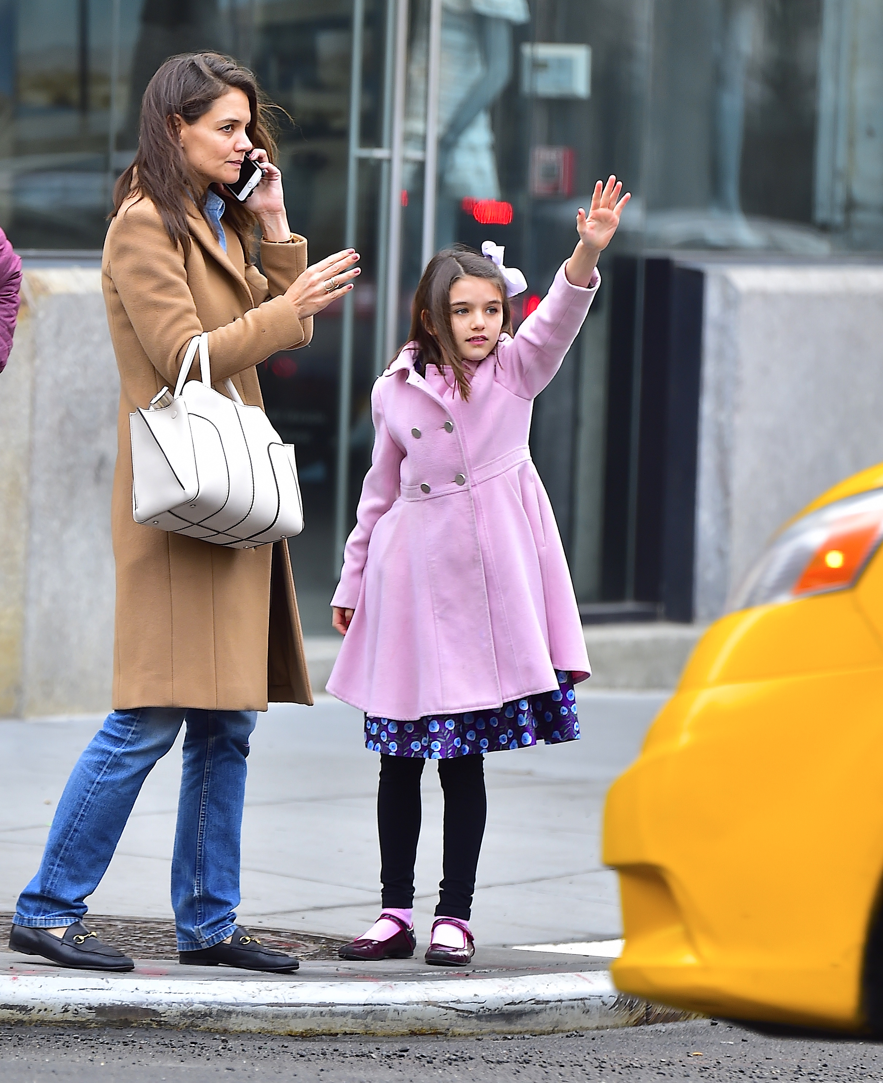 Katie Holmes stands gracefully on the curb, phone in hand, as her daughter Suri confidently hails a cab beside her. Wrapped in a stylish pink coat, Suri mirrors her mother’s poise with youthful flair. The scene captures a charming slice of city life filled with elegance, independence, and love in motion.
