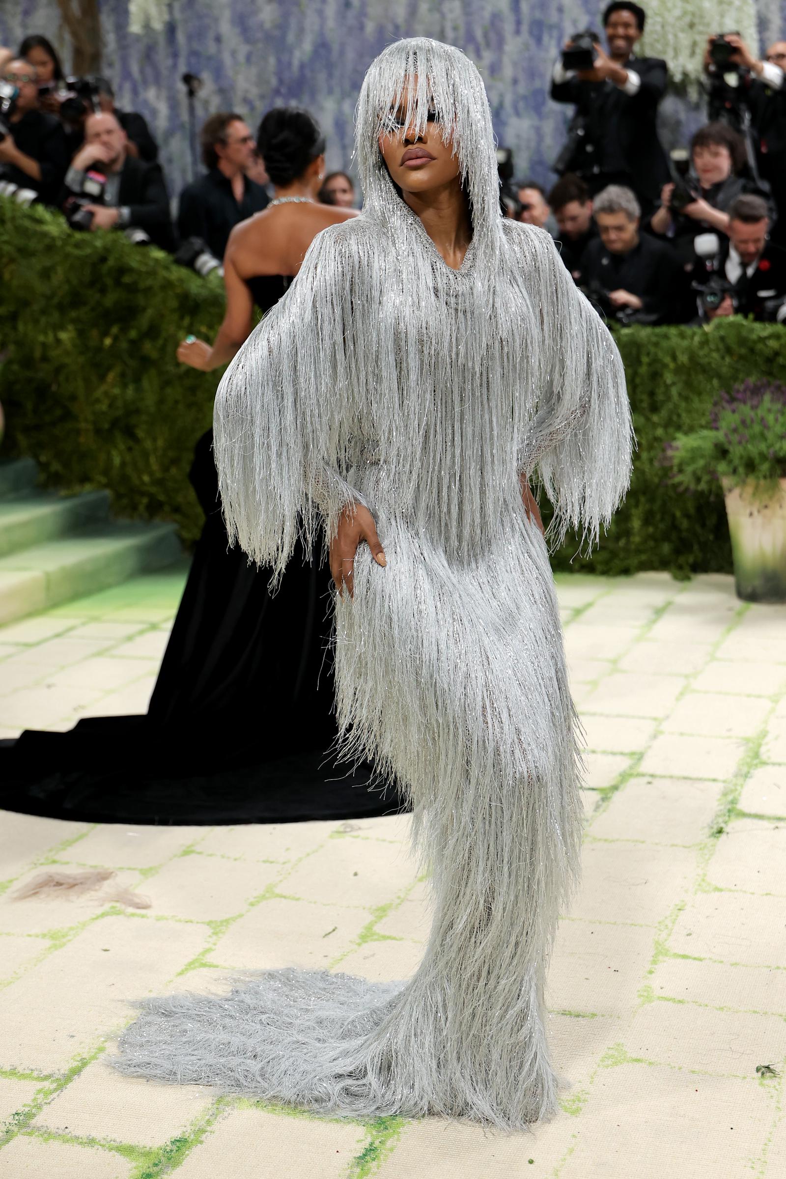 Teyana Taylor head-to-toe in a floor-length silver fringe gown, the dense metallic strands cascading over her face as a veil and continuing down oversized sleeves before pooling into a trailing hem.