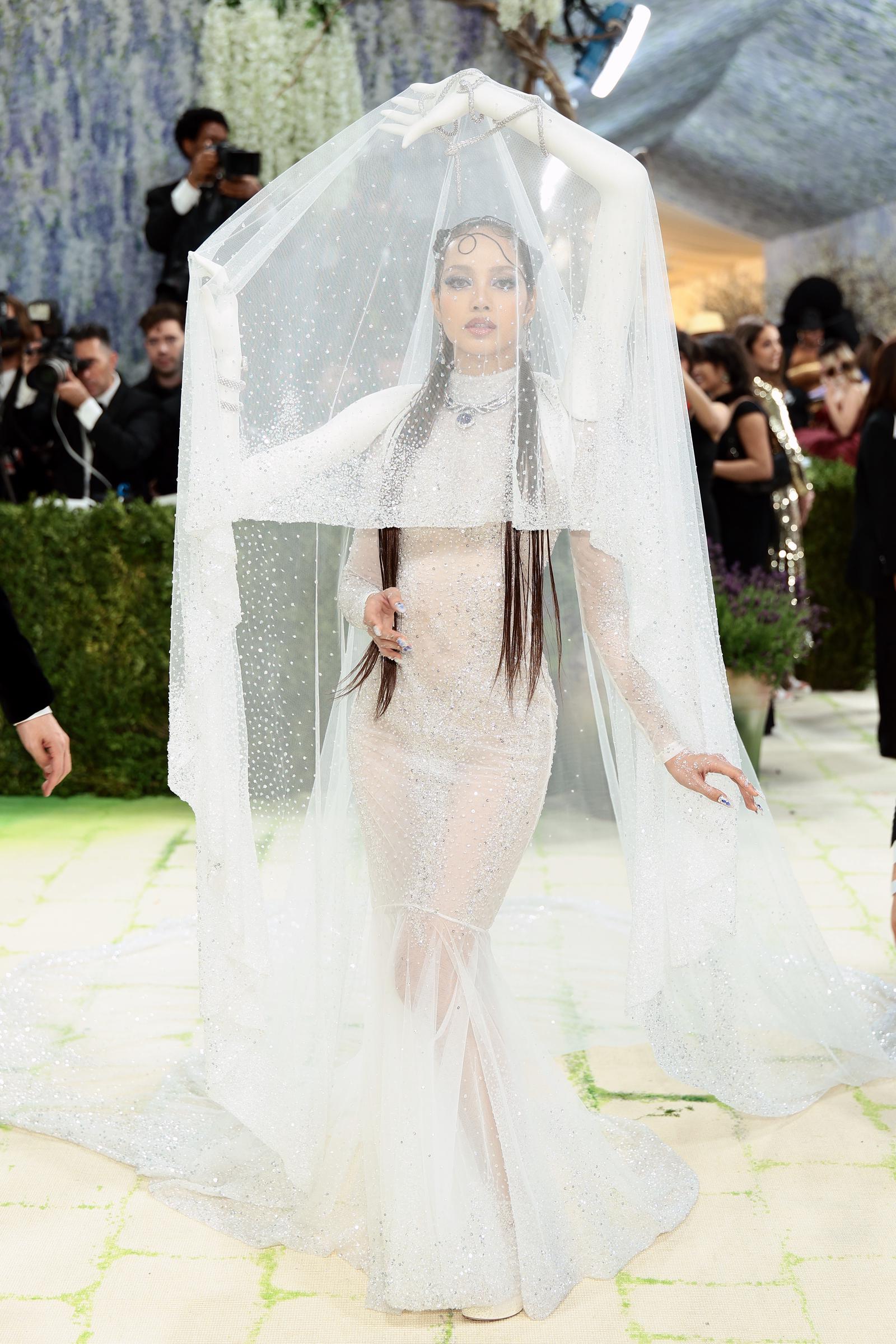 Lisa walks the carpet in a crystal-encrusted nude illusion fitted gown, its surface scattered with thousands of tiny stones, the sweeping sparkle-dotted tulle veil arching overhead and held aloft by ivory hands, while her own arms pose freely at her sides.